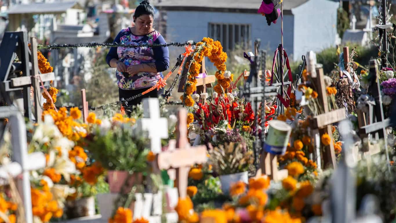 Una mujer está en un cementerio decorando una tumba con flores de cempasúchil y otras ofrendas.