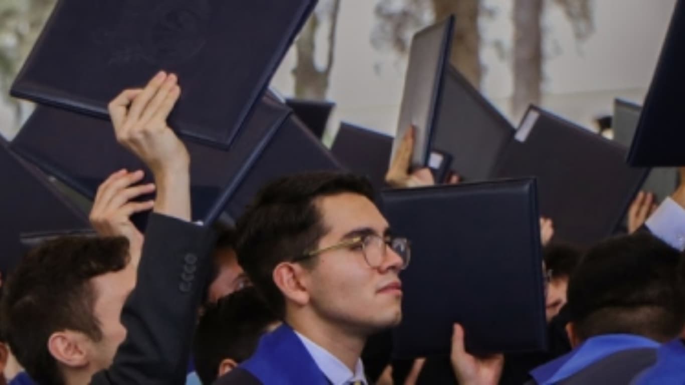 Un grupo de graduados sostiene sus diplomas en alto en una ceremonia de graduación. En primer plano, un joven con gafas se ve pensativo mientras sonríe. El fondo está lleno de otros graduados que celebran, creando un ambiente festivo y de logro