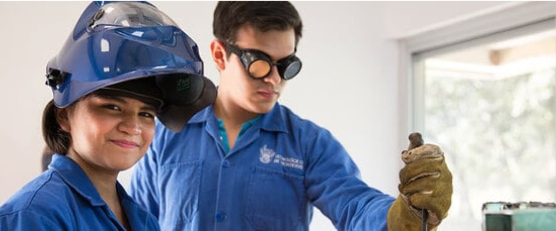 Una mujer y un hombre en un entorno de trabajo técnico, ambos vistiendo overoles azules. La mujer sonríe mientras usa un casco de seguridad azul, y el hombre, que lleva gafas de protección, sostiene un objeto con una mano enguantada. El fondo muestra una ventana con luz natural, sugiriendo un ambiente de aprendizaje práctico.