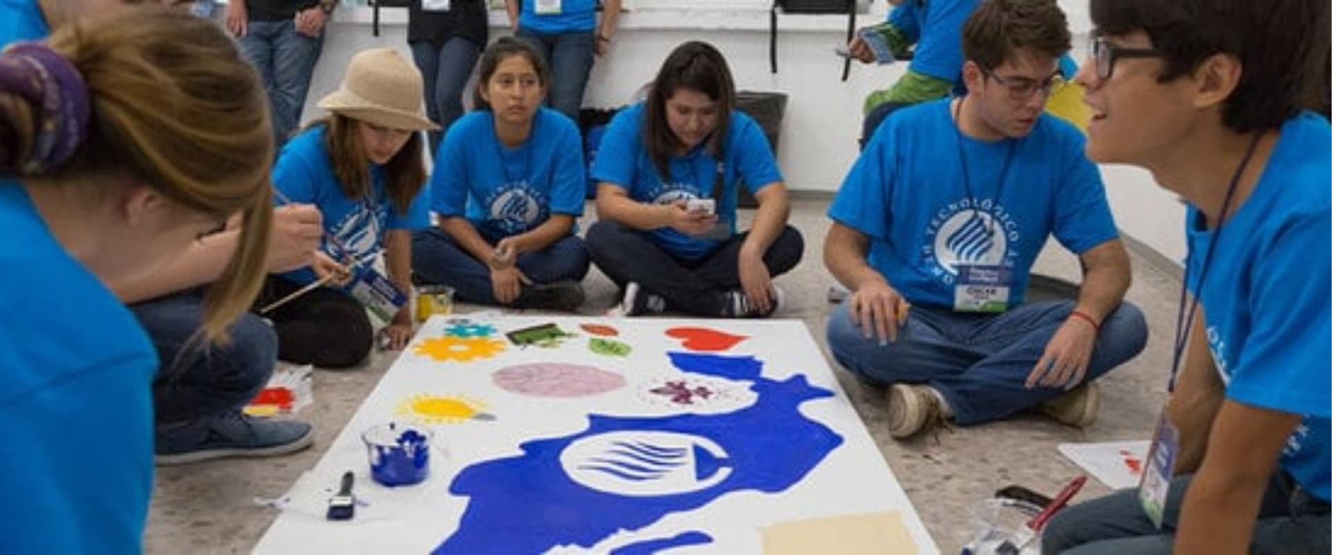 Un grupo de jóvenes vestidos con camisetas azules participa en una actividad creativa, pintando un gran mural en el suelo con colores vibrantes. Algunos están concentrados en el proceso artístico, mientras otros observan o usan sus teléfonos. El ambiente es colaborativo y lleno de energía creativa