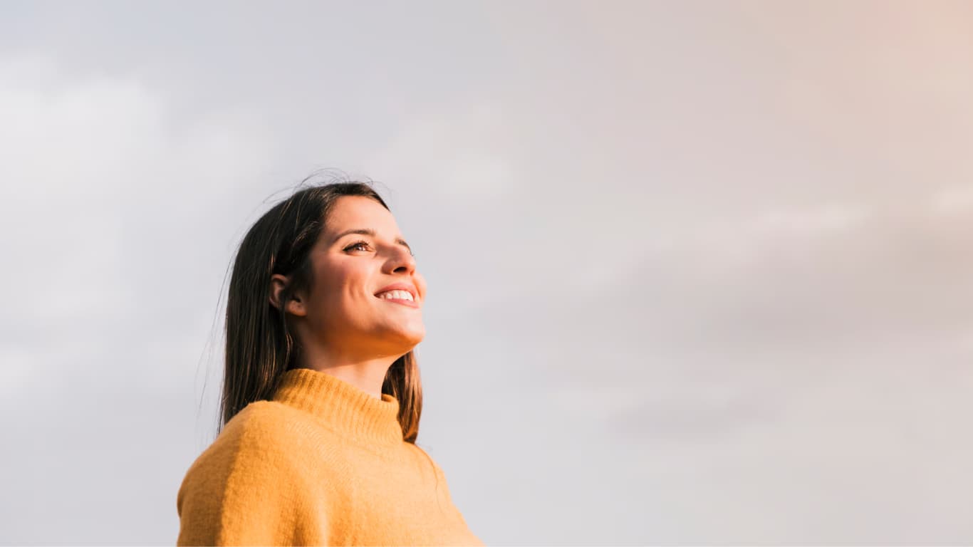 Una mujer joven con cabello lacio y largo sonríe mientras mira al horizonte. Lleva un suéter de color amarillo. El fondo muestra un cielo parcialmente nublado, creando un ambiente de paz y optimismo