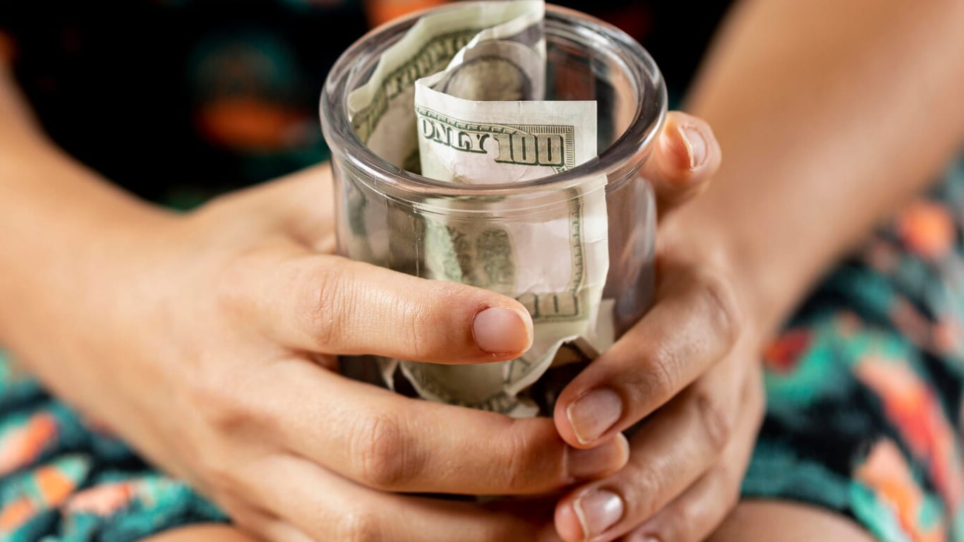 Una persona sostiene un frasco de vidrio lleno de billetes, mostrando solo las manos y el frasco.