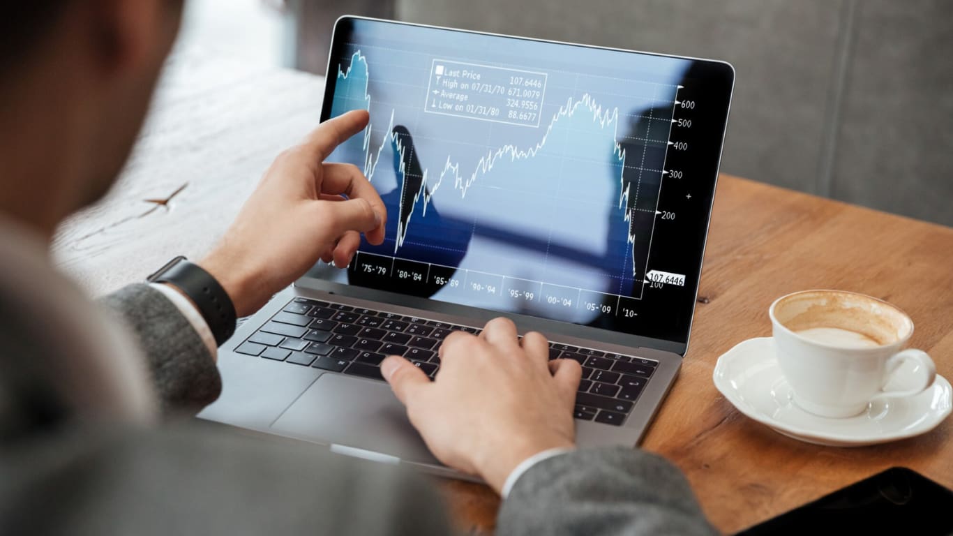 Un hombre está frente a una laptop, utilizando una gráfica de precios en la pantalla. Con una mano señala los datos, mientras que a su lado hay una taza de café.