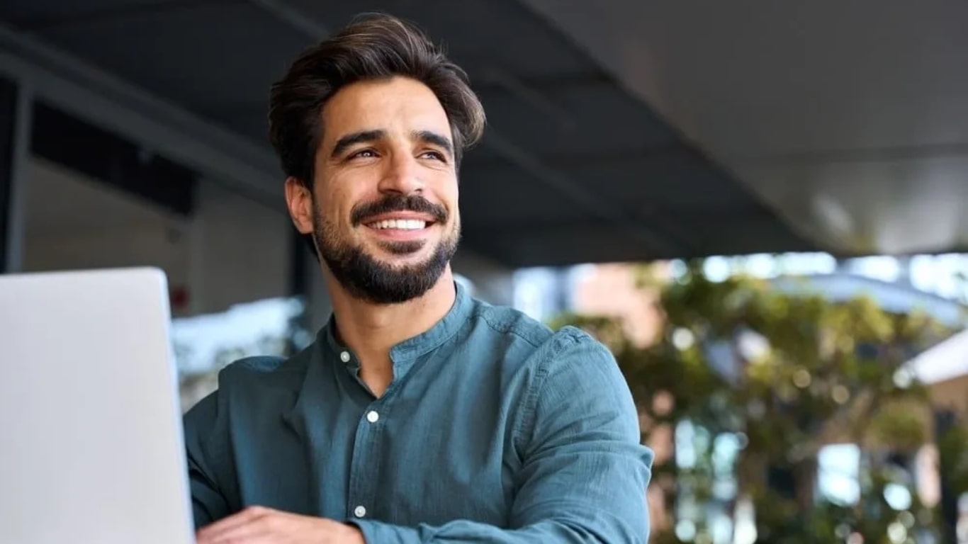 Un hombre sonriendo mientras trabaja en una computadora portátil al aire libre. Lleva una camisa de color verde y tiene barba.