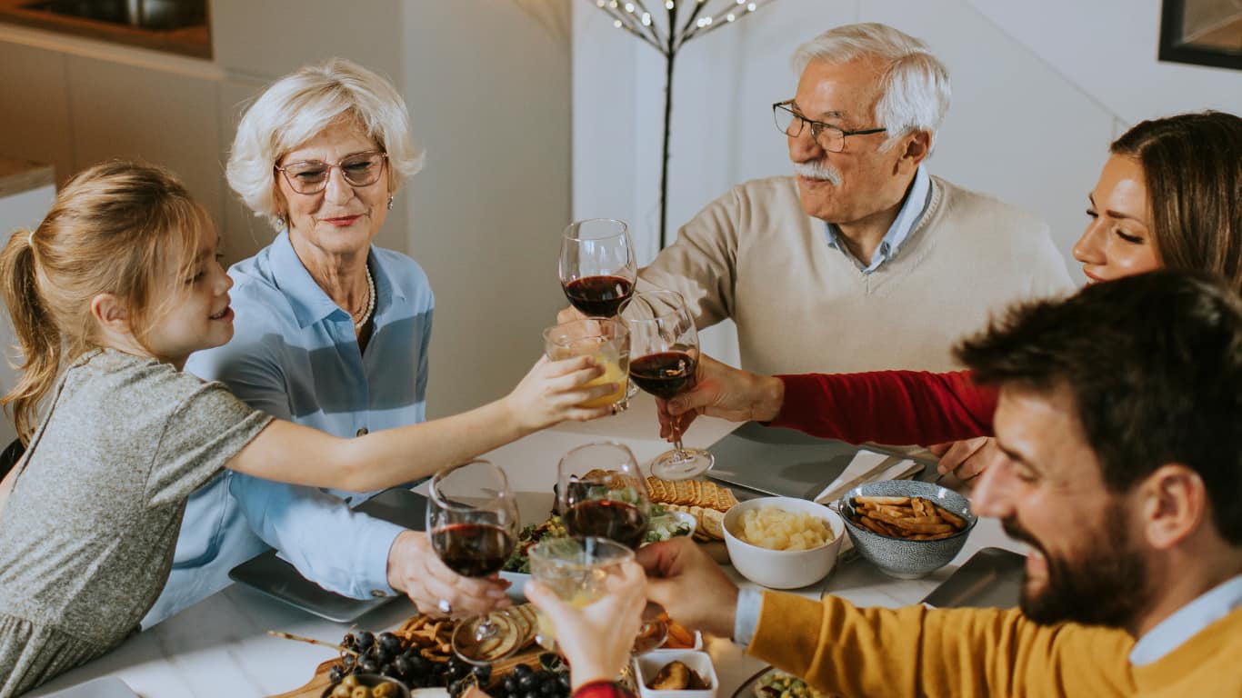 Un grupo de cinco personas, incluyendo una niña, celebra alrededor de una mesa repleta de comida y bebida. Todos levantan sus copas en un brindis, mostrando sonrisas y un ambiente festivo.