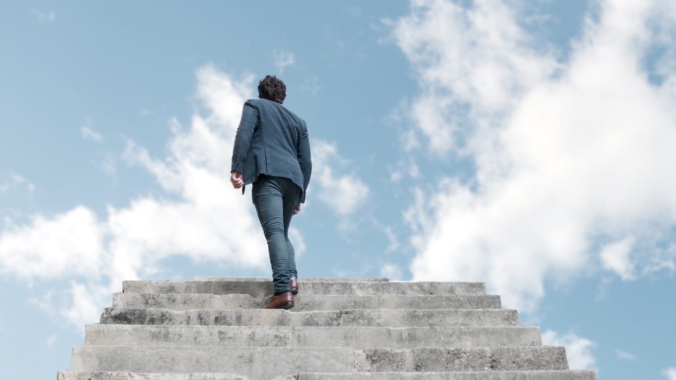 Un hombre con un traje oscuro camina hacia arriba por una serie de escalones de piedra, mirando hacia el cielo despejado con nubes esponjosas.
