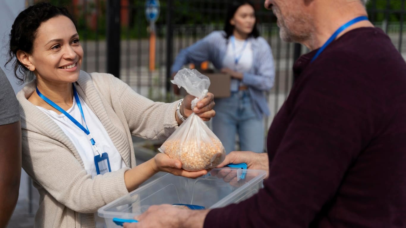 Una mujer sonriente, vistiendo un suéter claro y un gafete, está entregando una bolsa de alimentos a un hombre.