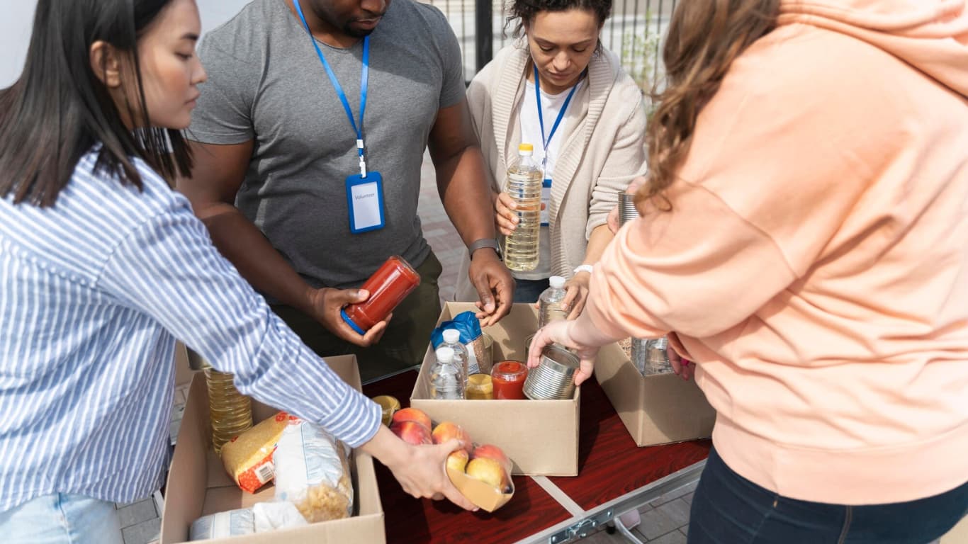 Un grupo de cuatro personas organizando alimentos y bebidas en cajas. Están concentrados en su tarea, manipulando latas, botellas y frutas.