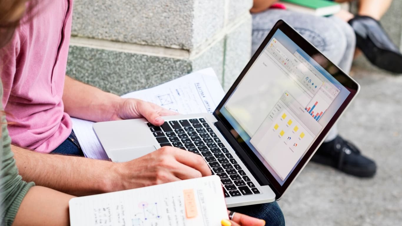 Una persona está sentada en el suelo, usando una laptop mientras sostiene hojas de papel. La pantalla de la laptop muestra gráficos y datos, mientras que la persona parece concentrada en su trabajo. 