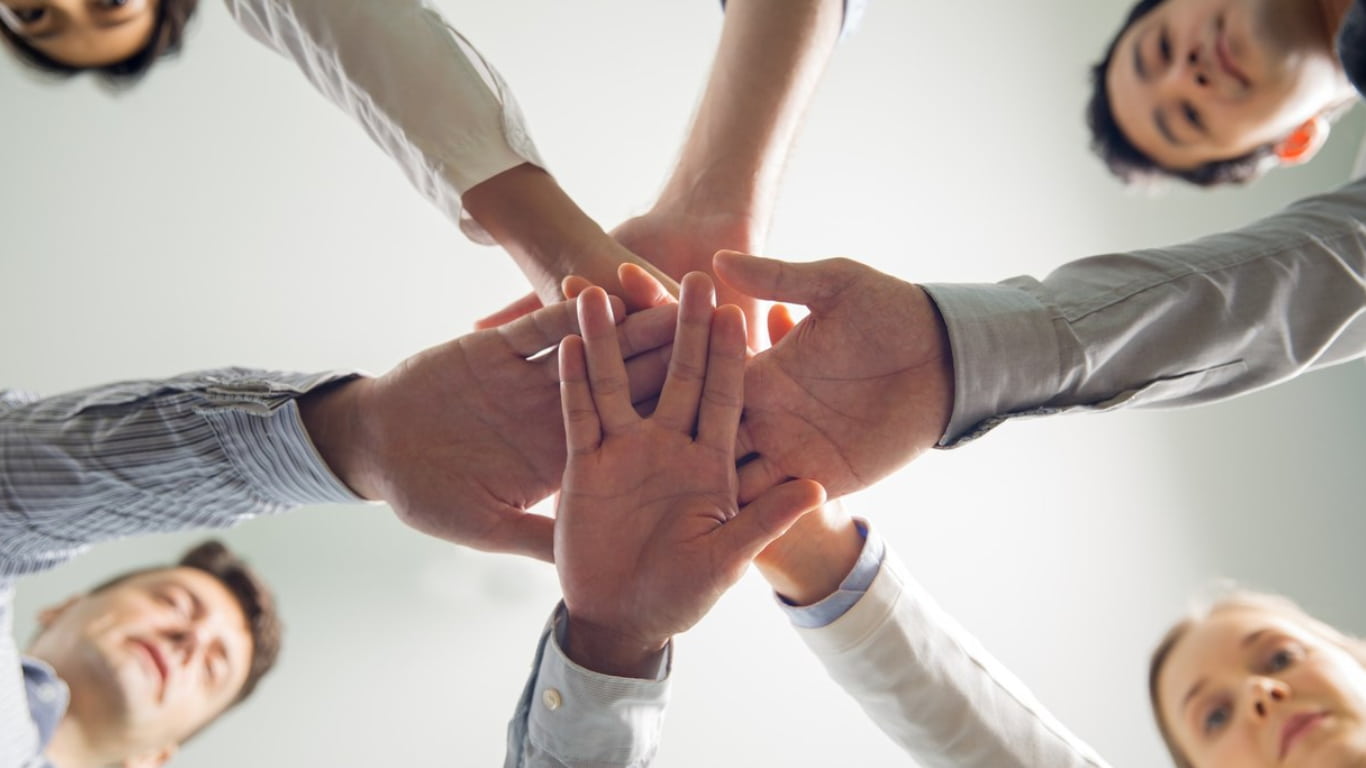 Un grupo de cinco personas está uniendo sus manos en el centro de la imagen, mostrando un gesto de colaboración y unidad.