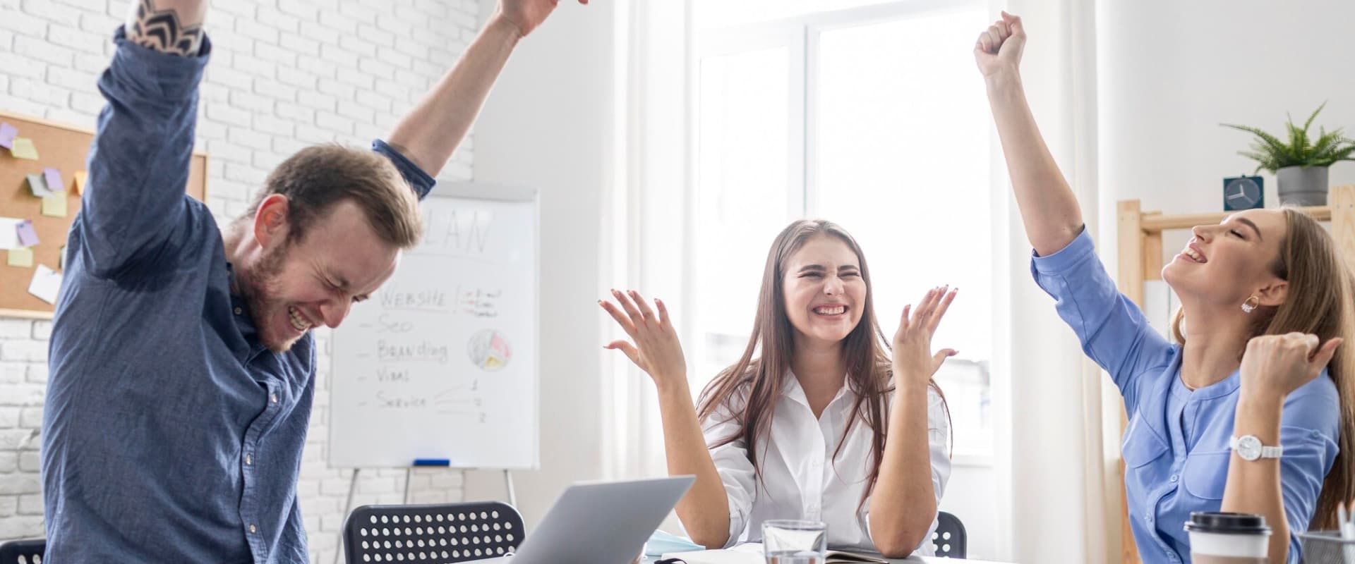 Hay una laptop y una pizarra blanca en el fondo, creando un ambiente de trabajo colaborativo y triunfante.