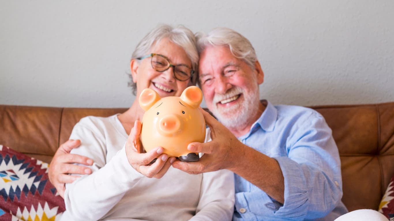 Una pareja de ancianos sonrientes está sentada en un sofá, sosteniendo una alcancía en forma de cerdito. Ambos lucen felices y están abrazados, disfrutando del momento.