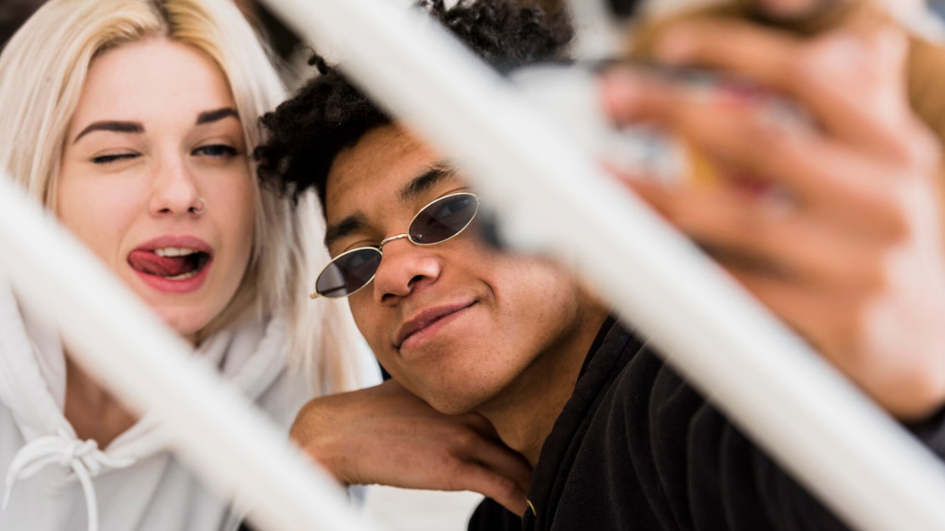 Dos jóvenes están posando para una selfie. La mujer tiene el cabello rubio y lleva una sudadera blanca, guiñando un ojo y sacando la lengua. El hombre tiene el cabello rizado y lleva gafas de sol, sonriendo a la cámara.