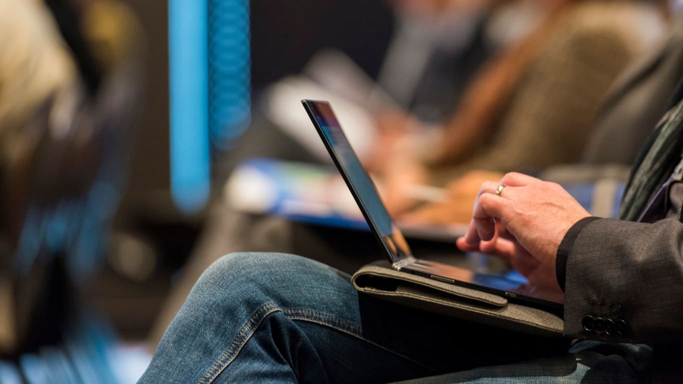 Un hombre está sentado en una conferencia, usando una laptop. Su mano se encuentra sobre el teclado, mientras que el fondo muestra a otras personas también prestando atención.