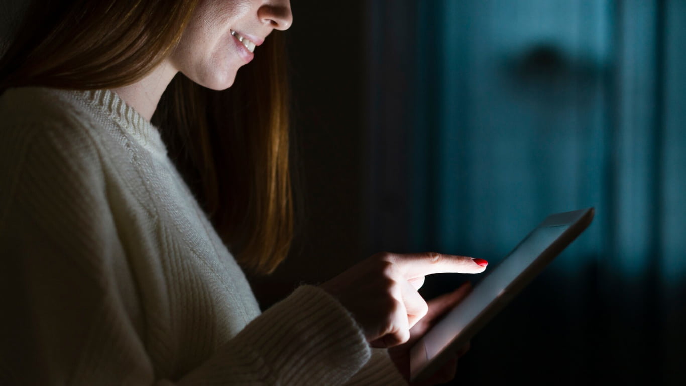 Una mujer está de pie en un ambiente oscuro, iluminando su rostro con una tablet que sostiene en sus manos.