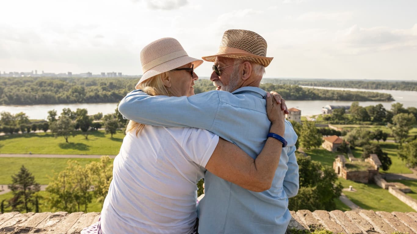 Una pareja de ancianos se abraza mientras contemplan un paisaje desde un alto. Ambos llevan sombreros y sonríen, disfrutando del momento juntos.