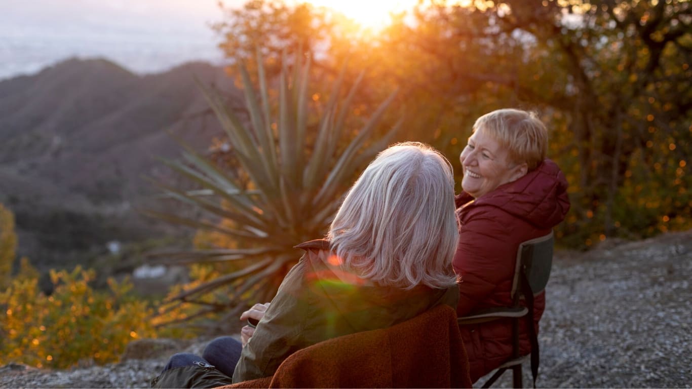 Dos mujeres mayores están sentadas en sillas al aire libre, disfrutando de la puesta de sol. Una de ellas tiene el cabello gris y lleva una chaqueta roja, mientras que la otra sonríe y parece relajada.