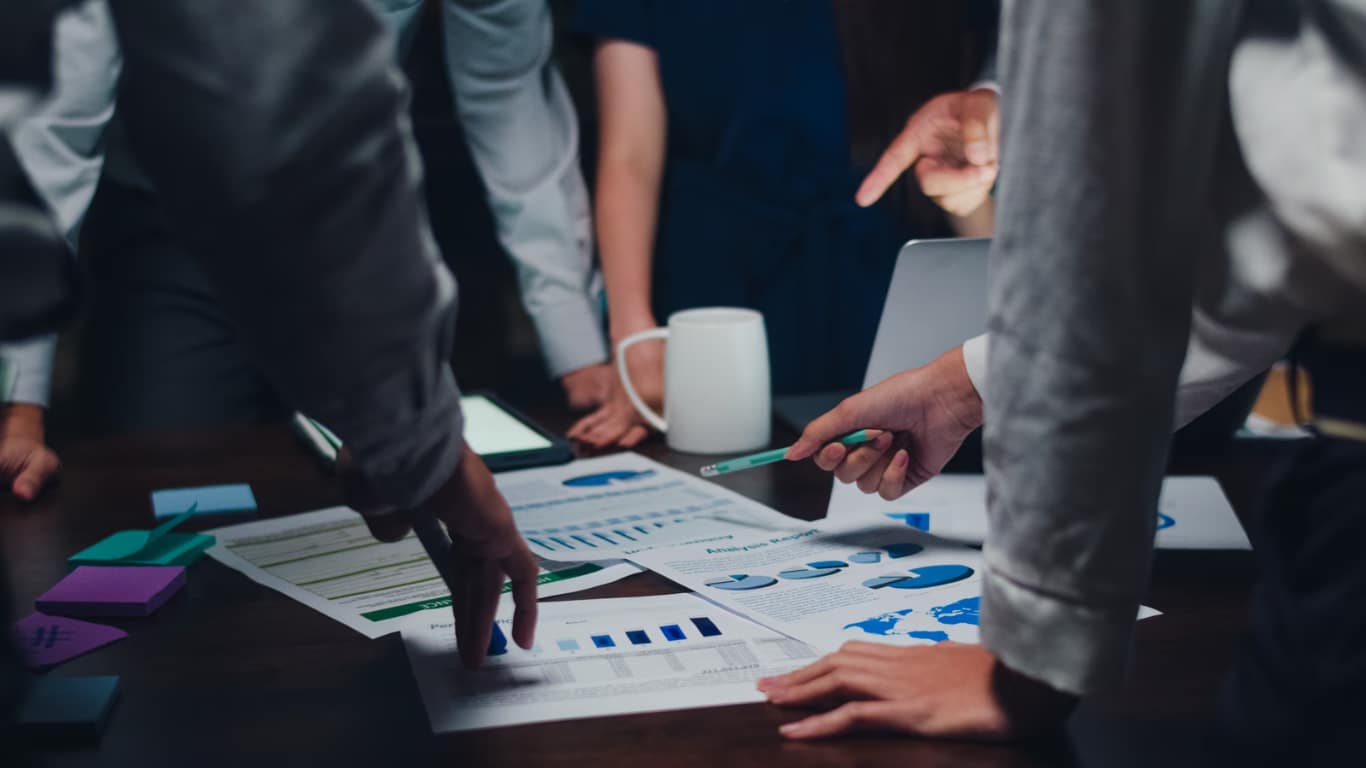 Varias manos están sobre una mesa, señalando gráficos y documentos relacionados con análisis de datos.