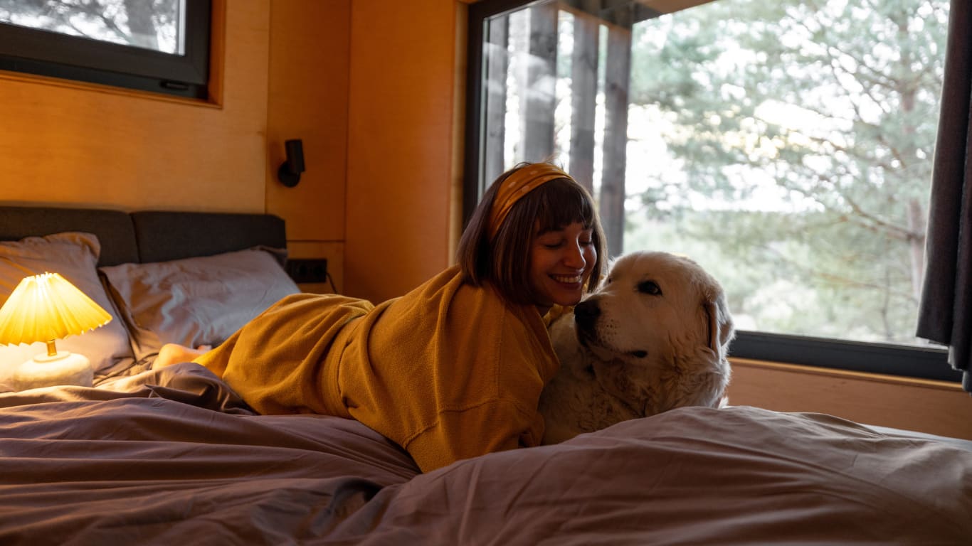 Una mujer sonriente se recuesta en una cama con sábanas grises, acariciando a un perro de raza golden retriever. La mujer lleva un suéter amarillo y una diadema a juego, mientras ambos miran hacia la cámara.