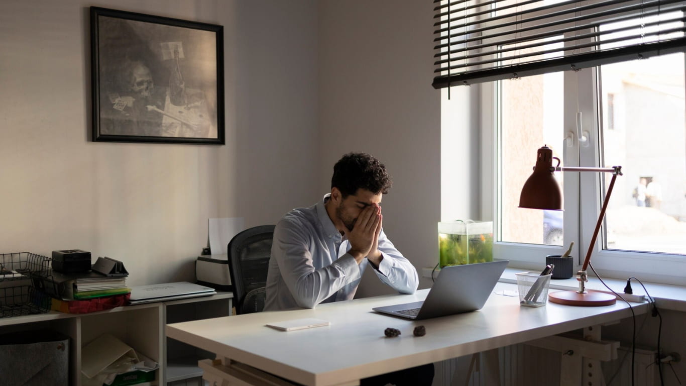 Un hombre sentado en un escritorio, con las manos en su rostro en un gesto de frustración o agotamiento.