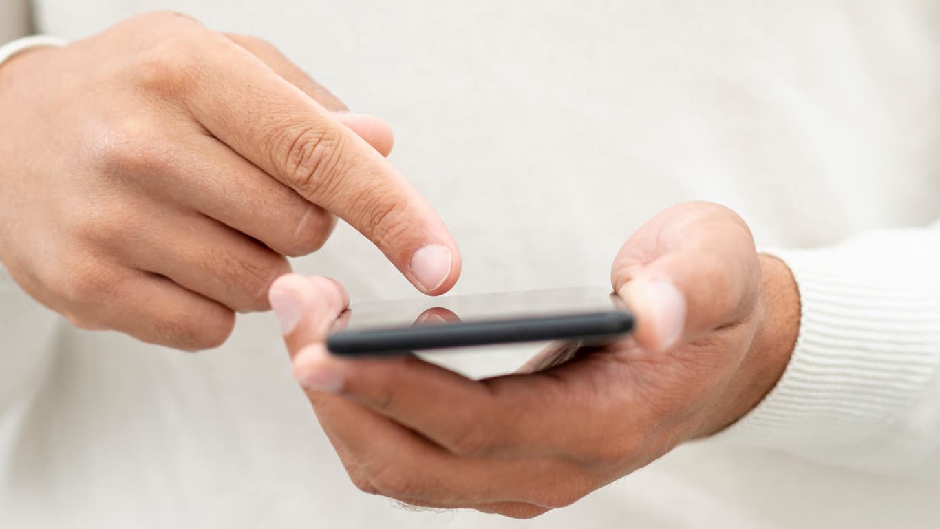 Una mano sostiene un teléfono inteligente mientras un dedo toca la pantalla. Del dispositivo emanan íconos brillantes de diversas aplicaciones, incluyendo un sobre, un carrito de compras, un corazón y un altavoz, simbolizando la comunicación y la conectividad digital. El fondo es suave y desenfocado, lo que resalta la acción en primer plano.