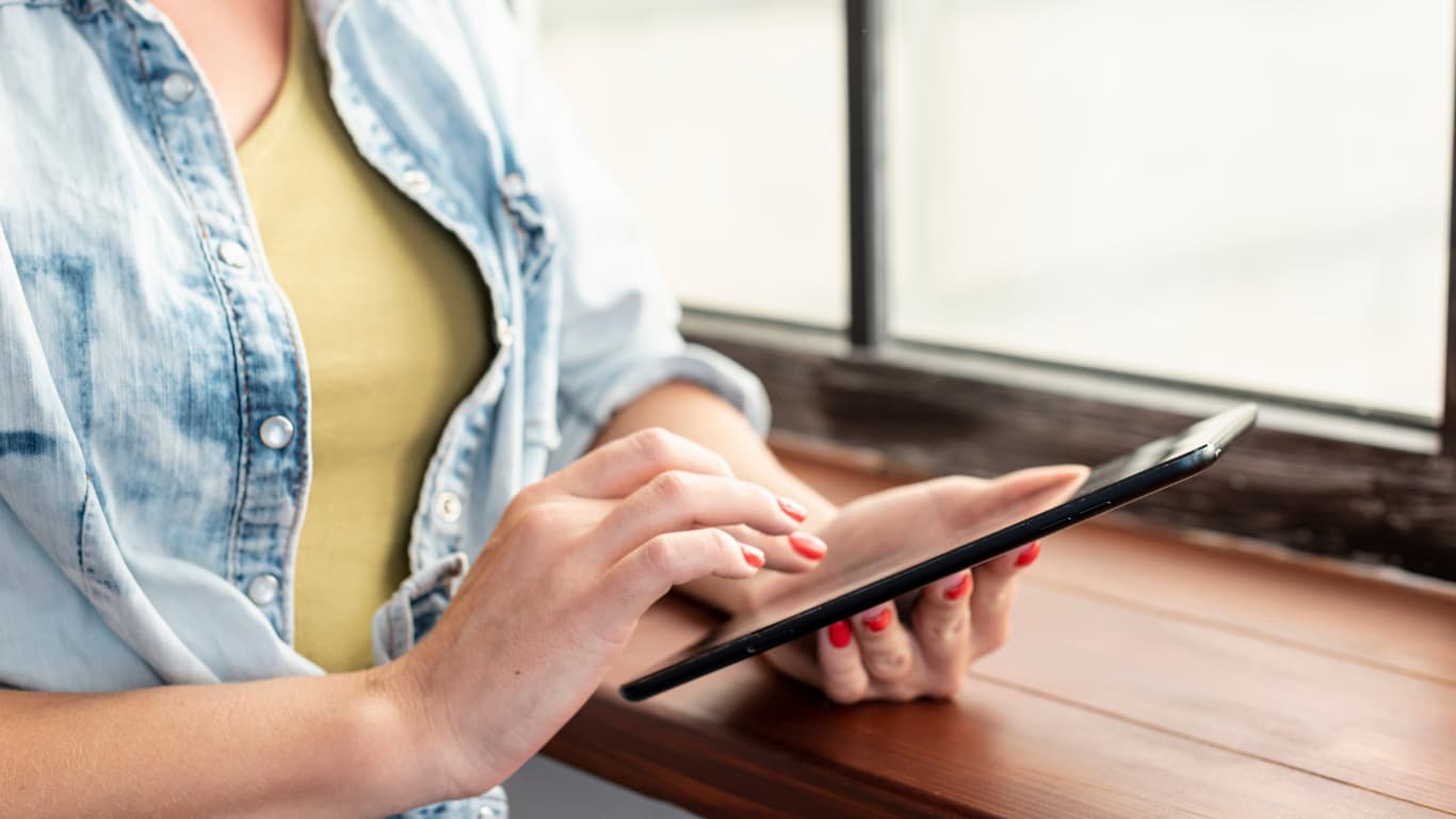 Una mujer sostiene una tablet mientras está sentada junto a una ventana. Lleva una camisa de mezclilla sobre una camiseta amarilla y muestra uñas pintadas de rojo. La luz natural entra por la ventana, iluminando suavemente el entorno y destacando la comodidad de la escena