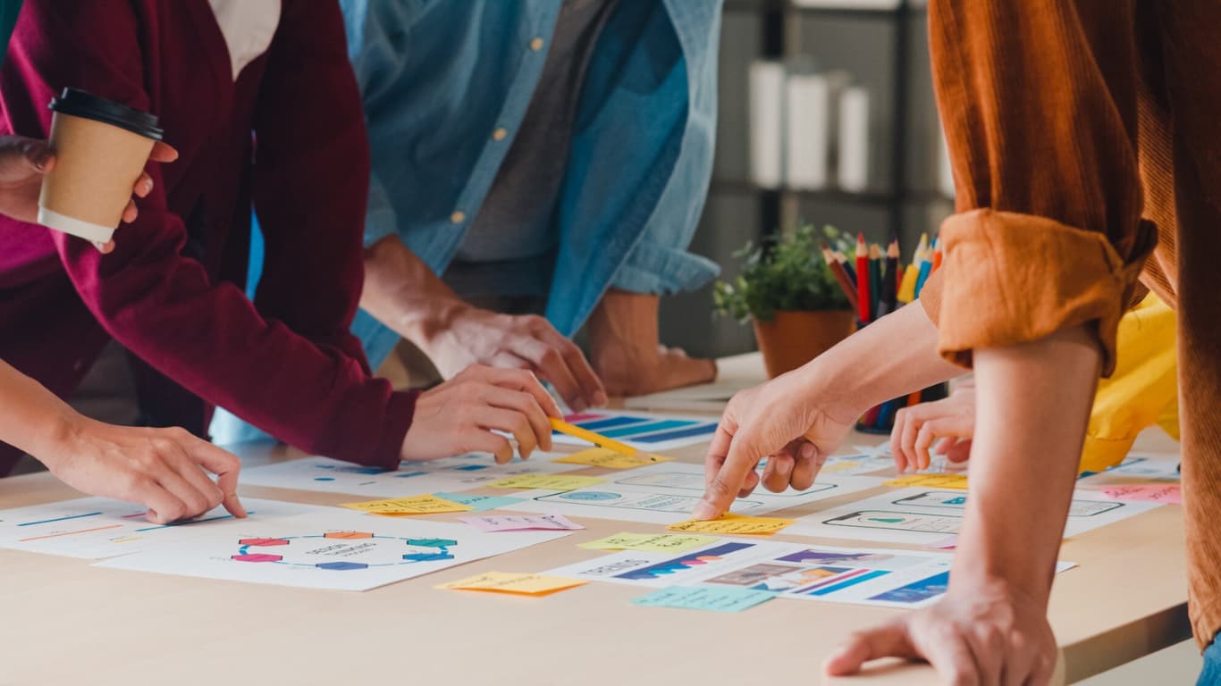 Un grupo de personas trabaja en una mesa, señalando gráficos y notas adhesivas de colores. Hay manos visibles que sostienen tazas de café y lápices, mientras interactúan en un ambiente colaborativo de trabajo, rodeados de documentos y un pequeño macetero.