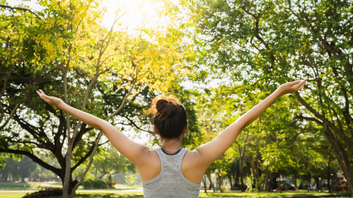 Una mujer de espaldas, con un peinado en moño y vestimenta deportiva, levanta los brazos hacia el cielo en un parque lleno de árboles. La luz del sol brilla a través de las hojas, creando un ambiente cálido y luminoso, simbolizando libertad y conexión con la naturaleza