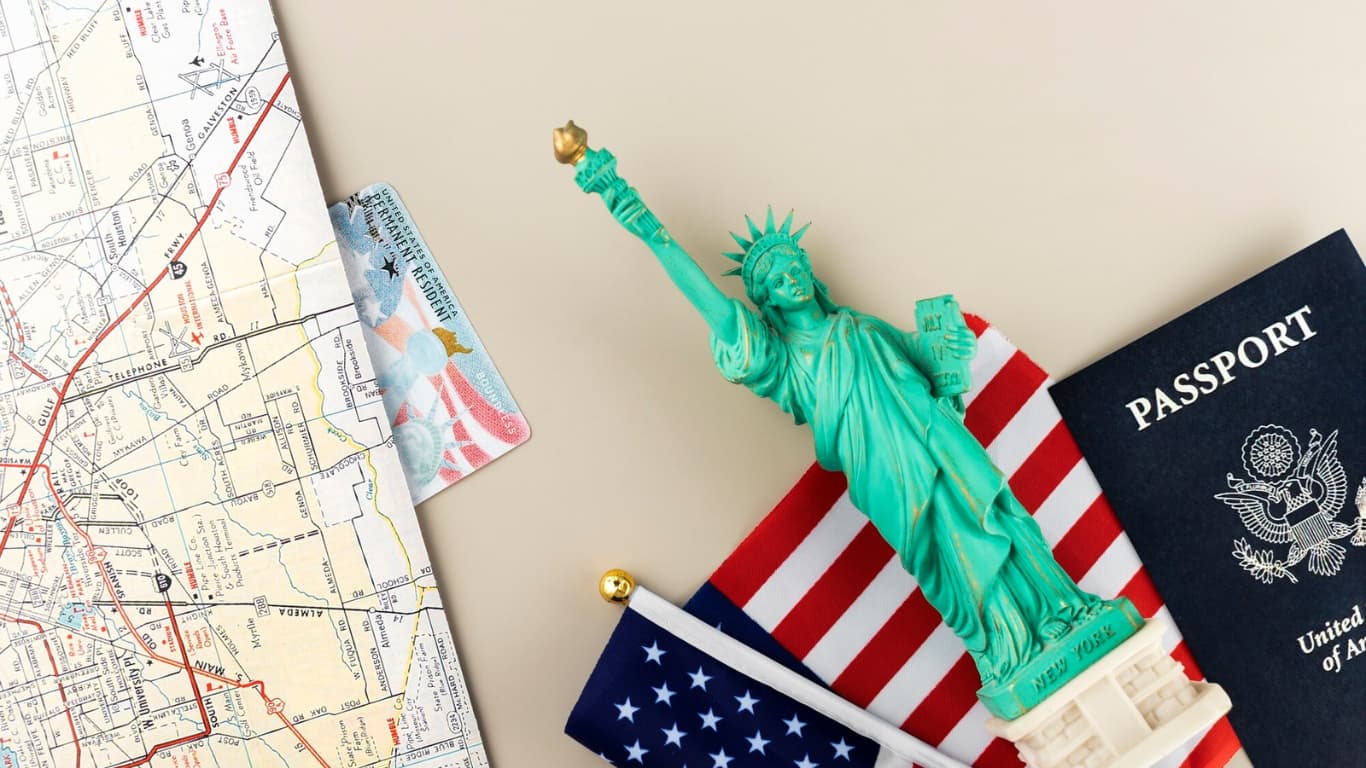 Una figura de la Estatua de la Libertad se encuentra sobre una mesa junto a un pasaporte estadounidense y una pequeña bandera de los Estados Unidos.