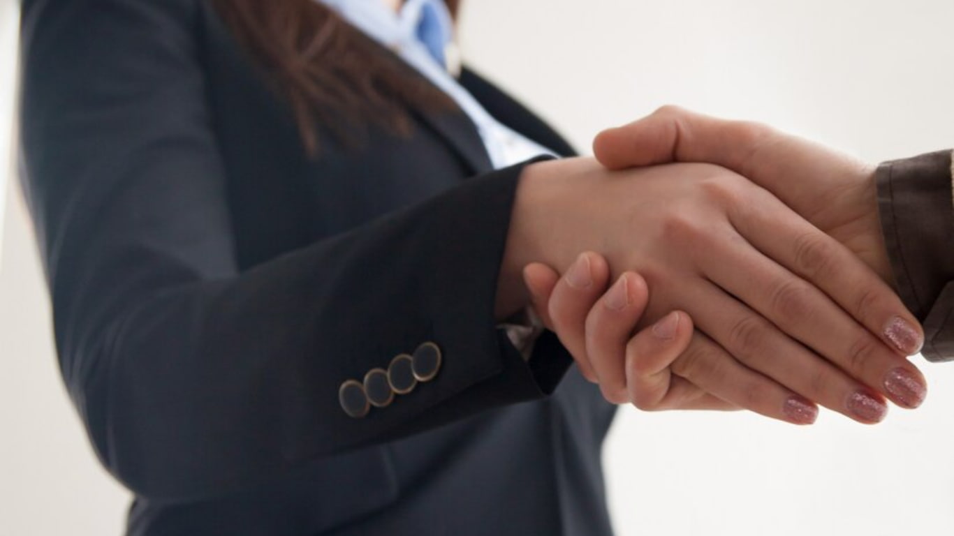 Dos manos están en un apretón de manos, simbolizando un acuerdo o un inicio de colaboración. La mano de la mujer está vestida con una chaqueta profesional, mientras que la otra mano es de un hombre, sugiriendo un contexto de negocios.