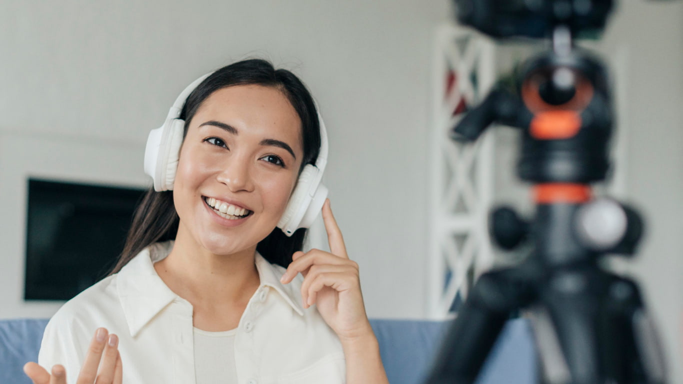 Una mujer sonriente lleva auriculares blancos y señala con un dedo mientras mira hacia una cámara montada en un trípode.