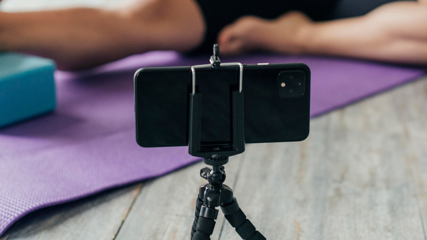 Un teléfono móvil está montado en un trípode, enfocando hacia una persona que está realizando ejercicios en una alfombra de yoga morada.