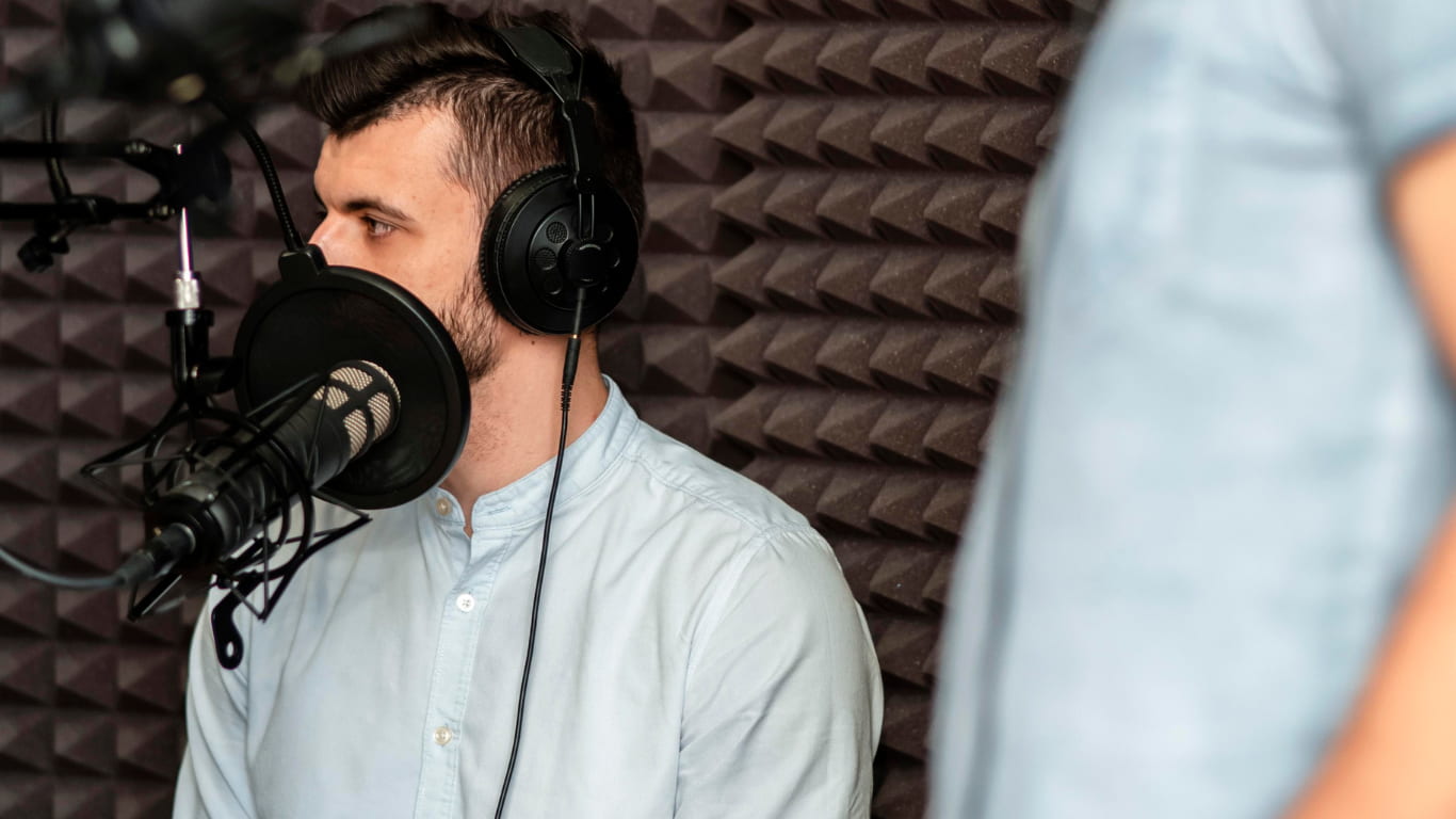 Dos hombres están en un estudio de grabación. Uno de ellos, sentado, lleva una camisa clara y auriculares, mientras habla frente a un micrófono. El otro, de pie y con una camiseta azul, observa.