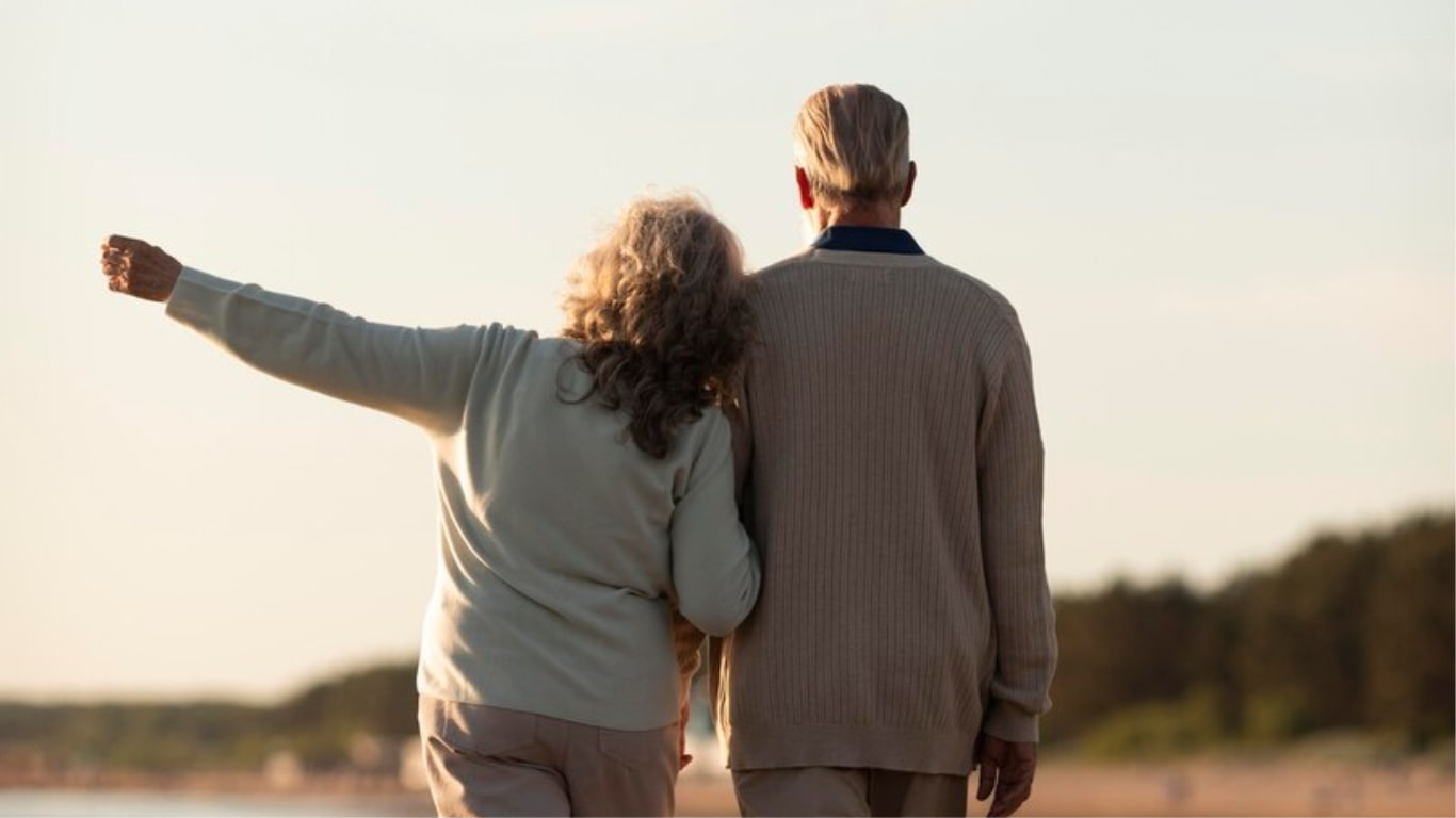 Una pareja se sienta en una manta en la playa, mirando hacia el horizonte al atardecer. La luz del sol brilla detrás de ellos, creando un aura dorada.
