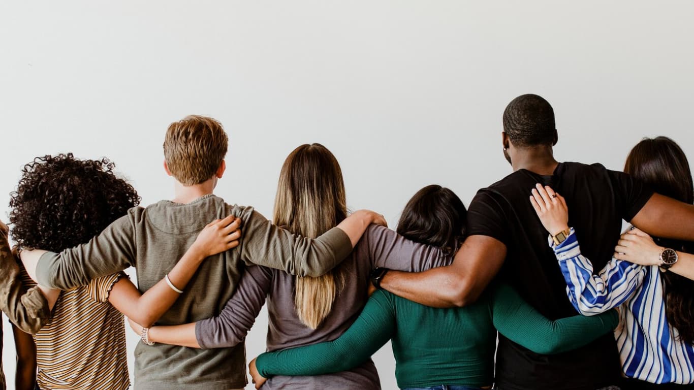 Vista posterior de un grupo diverso de personas de pie juntas, con los brazos alrededor de los hombros de los demás. La imagen muestra una variedad de estilos de vestimenta y tipos de cabello, reflejando unidad y camaradería