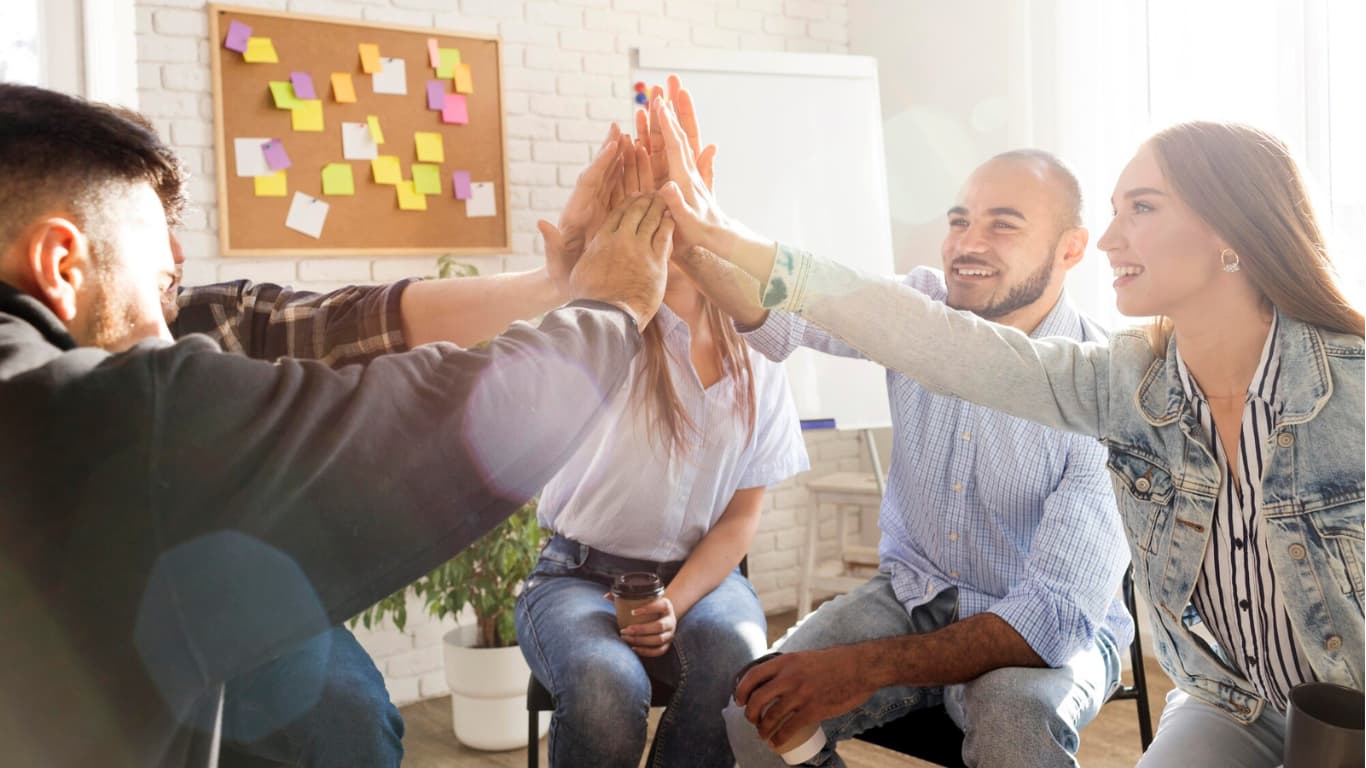 Un grupo de cinco jóvenes en un ambiente de oficina, realizando un gesto de 'high five' entre ellos. Todos lucen sonrientes y alegres, con algunas tazas de café en mano.