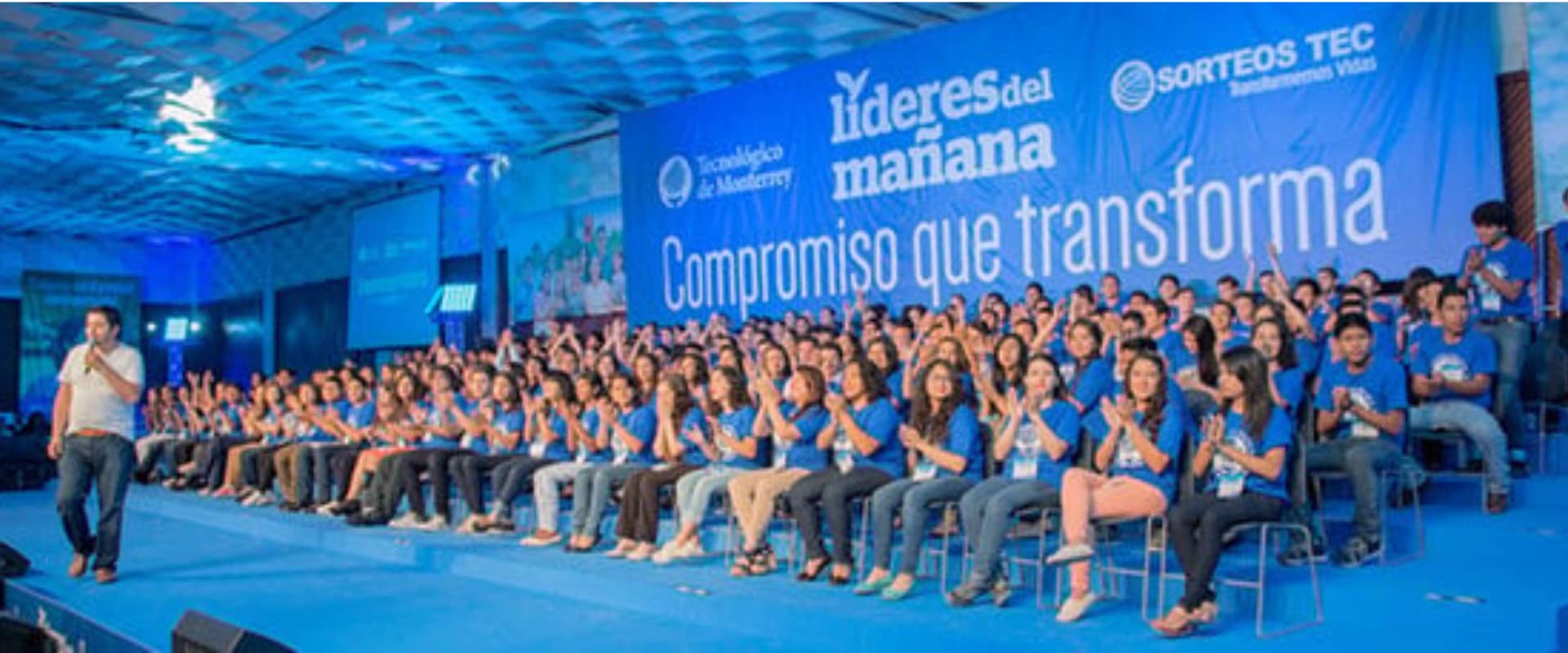 Una multitud de jóvenes aplaudiendo en un evento bajo un techo iluminado en tonos azules. En el escenario, un orador con micrófono se dirige a la audiencia. El fondo presenta un gran banner que dice 'Líderes del mañana' y 'Compromiso que transforma', creando un ambiente de energía y compromiso