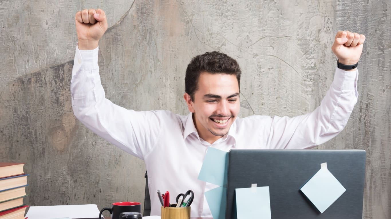 Un hombre sonriente levanta los brazos en señal de celebración frente a una computadora portátil en un escritorio.