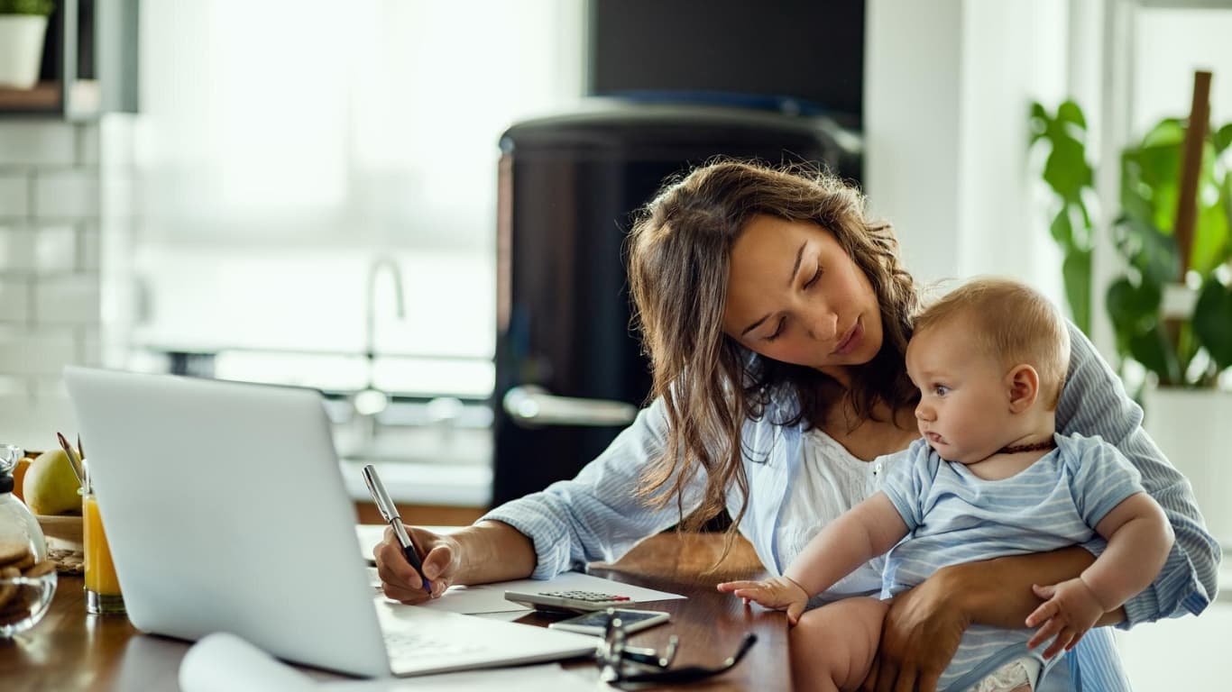 Una mujer está sentada en una cocina moderna, sosteniendo a un bebé en su brazo. Ella escribe en un cuaderno mientras mira a la computadora portátil frente a ella.