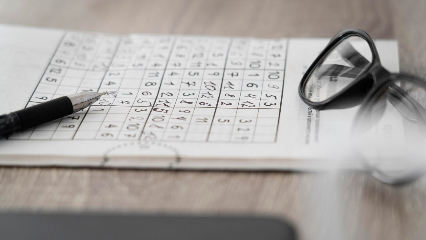 Una hoja de papel con un puzzle de sudoku en una mesa de madera, acompañada de un bolígrafo negro y unas gafas.