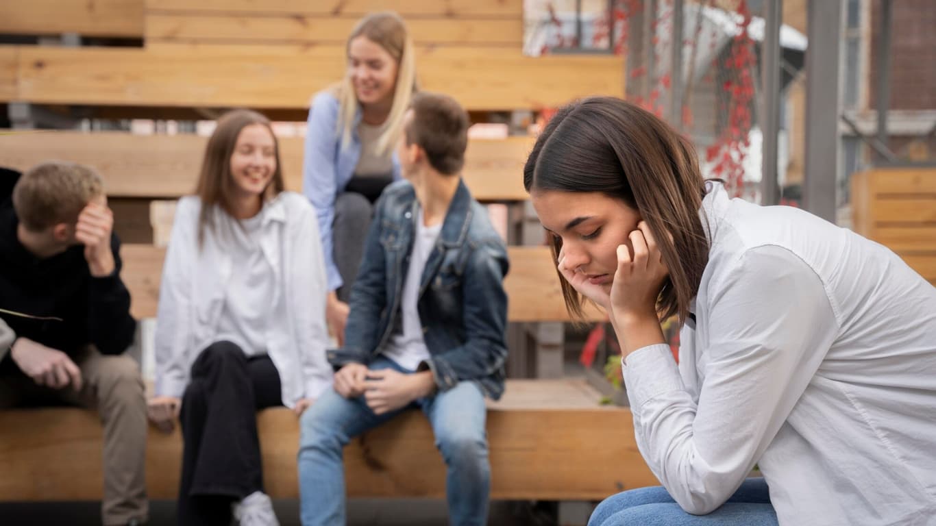 Un grupo de jóvenes sentados en una estructura de madera, charlando y riendo entre ellos. En primer plano, una mujer parece triste y reflexiva, con la cabeza inclinada y la mano en la mejilla, contrastando con la alegría del grupo.