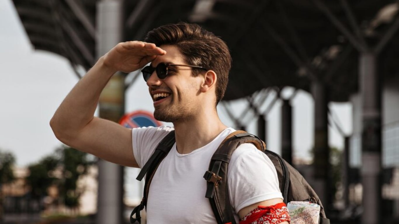Un hombre joven sonriente con gafas de sol y una camiseta blanca está de pie en una estación al aire libre, con una mano en la frente, como si estuviera buscando algo.