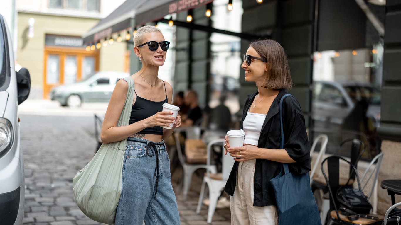 Dos mujeres jóvenes están de pie en una calle frente a una cafetería, conversando y sonriendo.