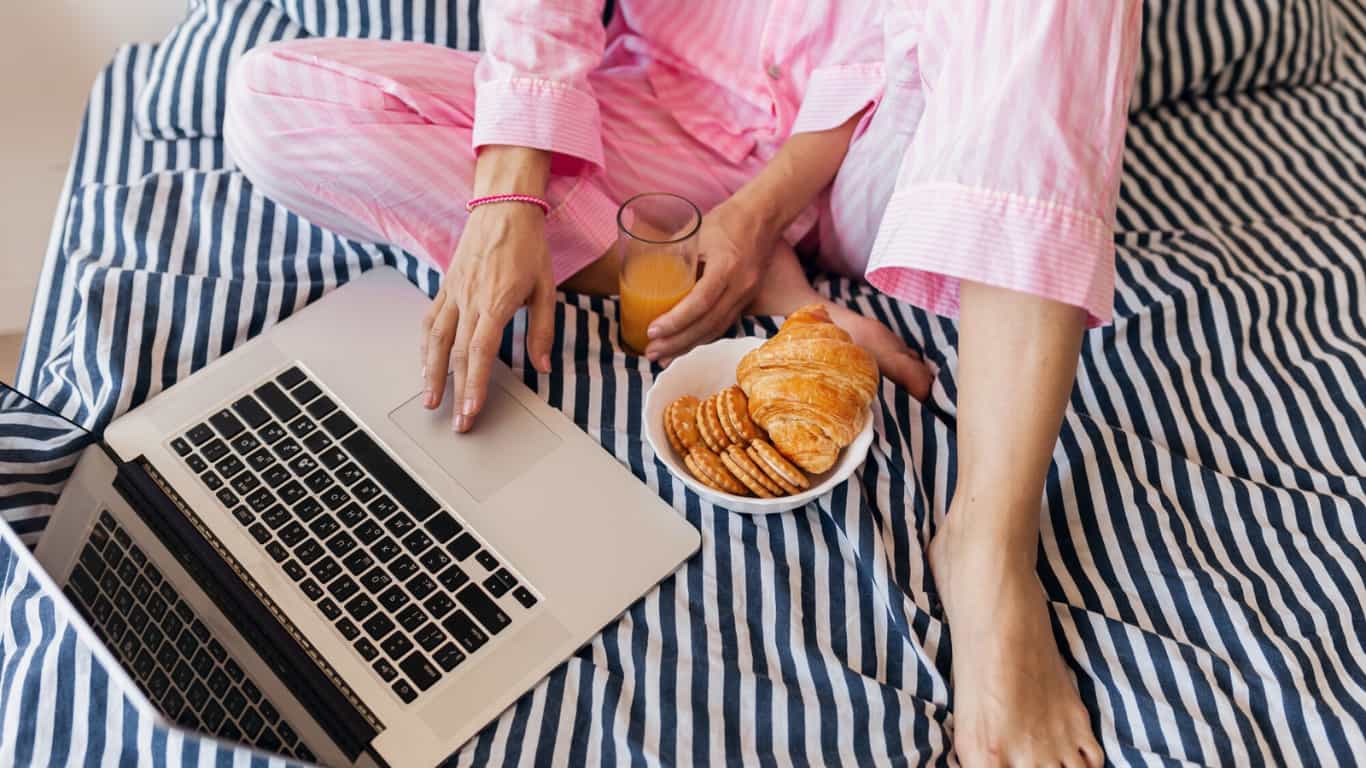 Una persona vestida con pijama de rayas rosas está sentada en la cama, usando una laptop. 