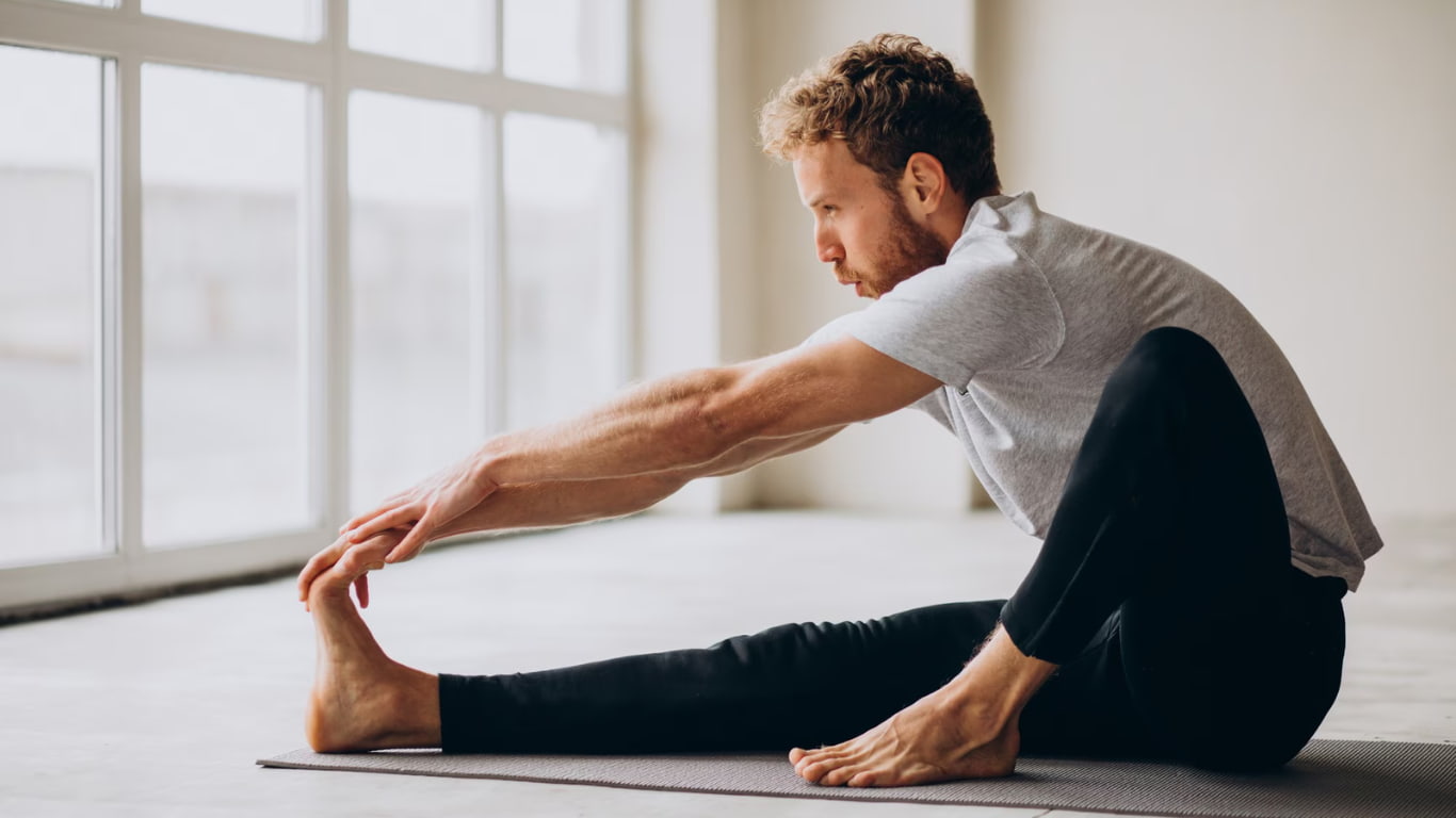 Un hombre está sentado en una esterilla de yoga, estirándose hacia adelante para alcanzar su pie. Viste una camiseta gris y pantalones negros, y el entorno es luminoso, con grandes ventanales que dejan entrar la luz natural.