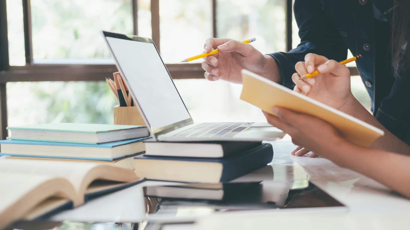 Dos personas colaboran en un escritorio, con una laptop abierta y varias pilas de libros. Una persona sostiene un cuaderno y un lápiz, mientras la otra apunta con un lápiz, sugiriendo un ambiente de estudio o trabajo en grupo.