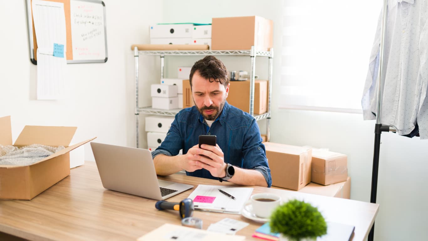 Un hombre con barba y cabello rizado, vestido con una camisa de mezclilla, mira su teléfono mientras está sentado en un escritorio. A su alrededor hay cajas de cartón y una laptop abierta. En la mesa también hay una planta pequeña y una taza de café, creando un ambiente de trabajo activo y organizado.