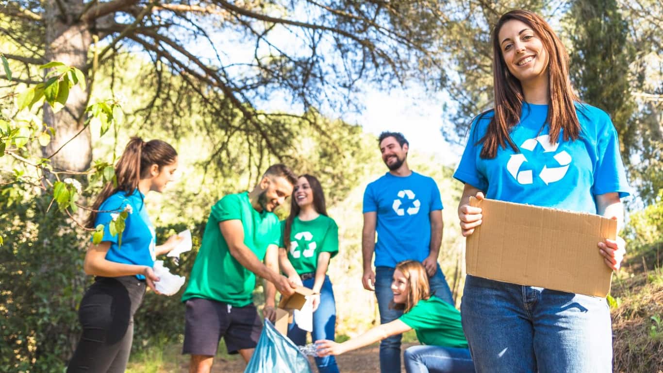 Un grupo de seis personas, vestidas con camisetas verdes y azules con el símbolo de reciclaje, están trabajando juntas en un entorno natural.