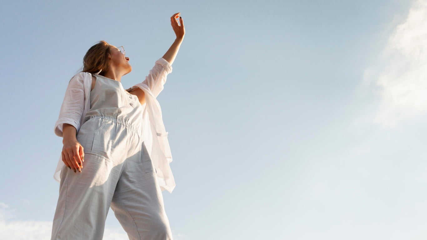 Una mujer de cabello largo y rubio está de pie, levantando un brazo hacia el cielo. Lleva un overol claro y una camisa blanca, y se encuentra bajo un cielo despejado.