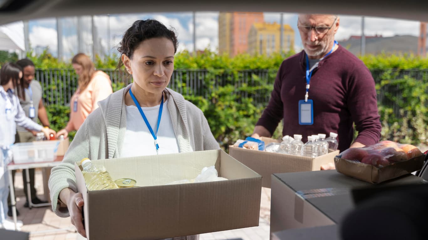 Una mujer, identificada como voluntaria por su etiqueta, sostiene una caja con productos, incluido aceite y otros artículos, mientras mira hacia adelante con concentración. Detrás de ella, un hombre se prepara para cargar otra caja con botellas de agua y alimentos.